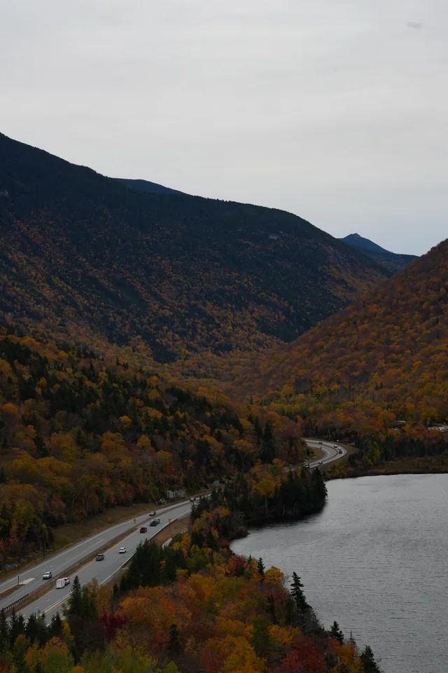 A lake and curving highway below an autumn-covered mountain.
